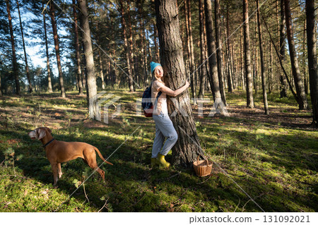 Relaxed woman hugging tree trunk in autumn forest with closed eyes at sunset, walk with dog in woods Relaxed woman hugging tree trunk in autumn forest with closed eyes at sunset, walk with dog in woods 131092021