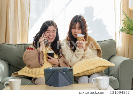 Winter day brings women friends together at home, enjoying cozy friendship indoors, talking on phone, watching tablet with warm coffee, embracing comforting winter leisure activities. 131094832