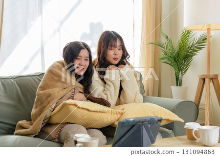 Winter home friends cherish cozy friendship, two smiling women enjoy watching digital device on sofa, warm blankets and hot drinks provide comfort during brisk winter weather at home. 131094863