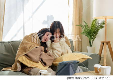 Winter day at home, women friends enjoy cozy friendship, sharing warm blankets, watching digital tablet content, creating happy moments indoors during cold winter season, relaxing comfort. 131094868