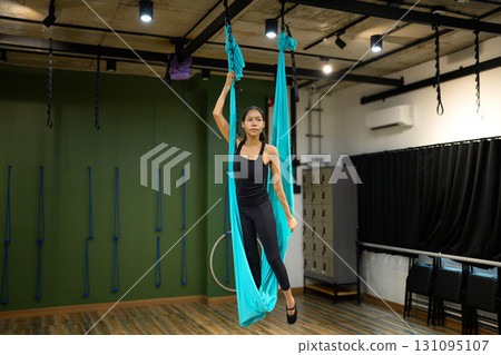 Female athlete standing confidently with aerial yoga swing, focusing on strength and balance training 131095107