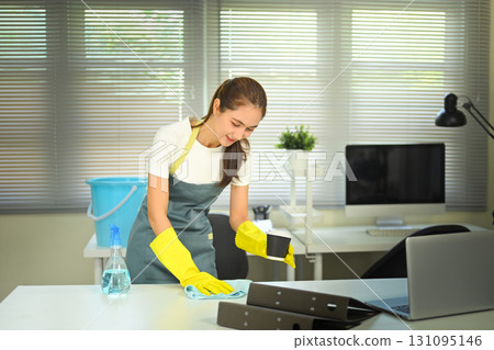 Smiling woman in apron and yellow gloves cleaning office desk with cloth and spray bottle Smiling woman in apron and yellow gloves cleaning office desk with cloth and spray bottle 131095146