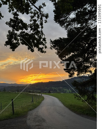 A stunning sunset paints brilliant colors over a serene landscape with trees and a winding road 131095691