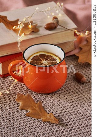 Cozy autumn still life with an orange mug of tea, books, dry leaves and warm decorative lights 131096818