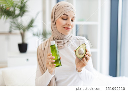Young Muslim woman holding avocado and lotion bottle indoors, promoting natural skincare routine 131099210