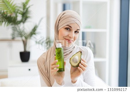 Young Muslim woman in beige hijab holding half avocado and skincare lotion promoting natural beauty and skincare. Indoor setting with serene and natural ambiance. Young Muslim woman in beige hijab holding half avocado and skincare lotion promoting natural beauty and skincare. Indoor setting with serene and natural ambiance. 131099217