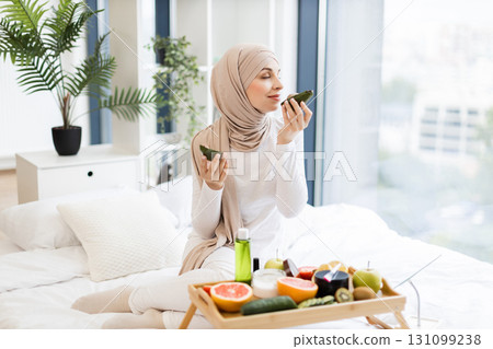 Young Muslim woman in modern living space using avocado and fruits for natural homemade skincare treatment. Relaxation and self-care activity while promoting healthy lifestyle and personal wellness. 131099238