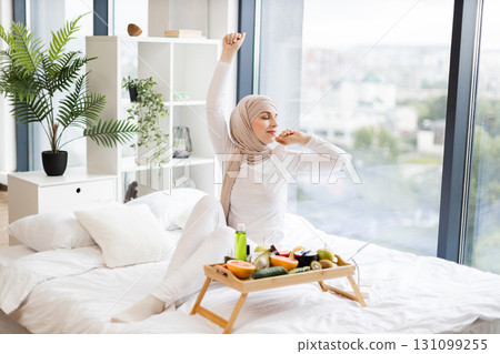 Young adult Muslim woman in modern interior sitting on bed preparing for skincare using fresh natural ingredients placed on table. Bright and welcoming atmosphere captured in cozy home environment. Young adult Muslim woman in modern interior sitting on bed preparing for skincare using fresh natural ingredients placed on table. Bright and welcoming atmosphere captured in cozy home environment. 131099255