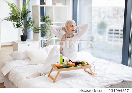 Young Muslim woman enjoying fresh morning in bedroom with healthy fruit breakfast tray. Relaxing atmosphere conveys health and calmness. The room is modern, bright, and adorned with greenery. 131099257