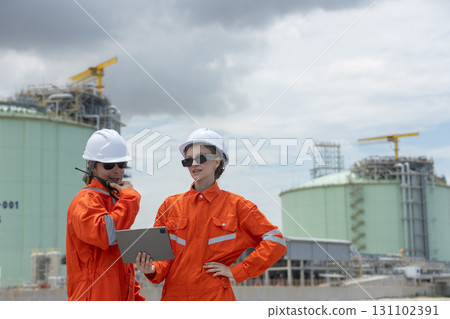 Two engineers in orange uniforms and safety helmets discussing project data at a gas plant with digital tablet and radio, representing teamwork, technology, energy industry, and field operations. Two engineers in orange uniforms and safety helmets discussing project data at a gas plant with digital tablet and radio, representing teamwork, technology, energy industry, and field operations. 131102391