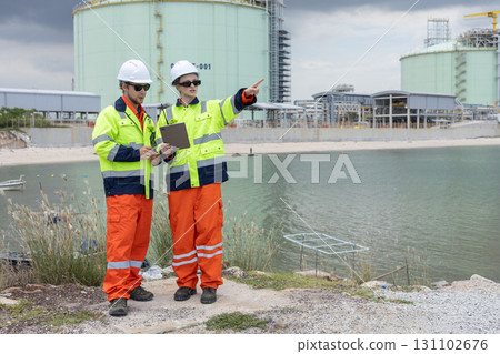 Two engineers wearing high-visibility safety jackets and helmets standing by the water, discussing inspection plans and pointing toward a large industrial gas storage facility in the background. Two engineers wearing high-visibility safety jackets and helmets standing by the water, discussing inspection plans and pointing toward a large industrial gas storage facility in the background. 131102676