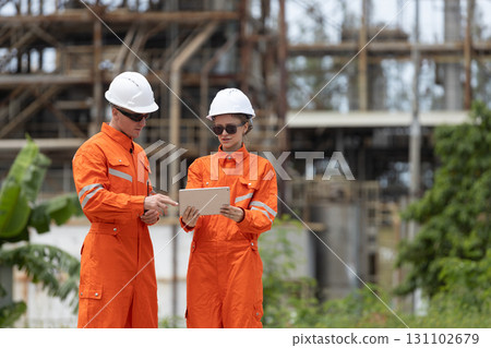 Two engineers in orange safety uniforms and white helmets reviewing project details on a tablet at an industrial construction site, representing teamwork, digital planning and field coordination. Two engineers in orange safety uniforms and white helmets reviewing project details on a tablet at an industrial construction site, representing teamwork, digital planning and field coordination. 131102679