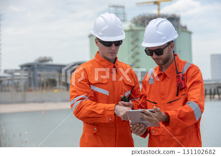 Two engineers in orange safety uniforms and helmets using a digital tablet to review field operations at a gas or energy facility, showing teamwork, communication and industrial inspection. 131102682