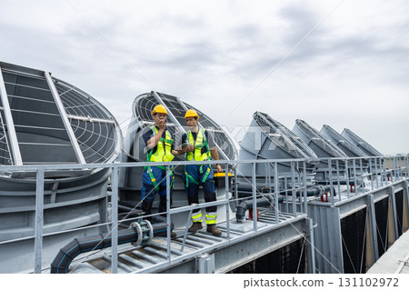 Two engineers in safety gear inspect rooftop cooling tower systems for building maintenance and energy optimization. Concept of HVAC technology, facility management, and industrial operation. 131102972