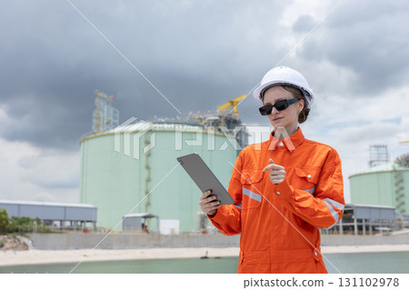 Female engineer in orange safety uniform and white helmet using a tablet while inspecting an industrial gas storage facility, representing technology, safety, and energy industry operations. 131102978