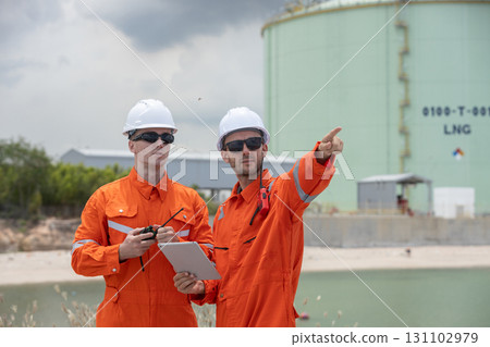 Two male engineer in orange protective uniforms and white helmets discussing site operations at LNG gas plant with a tablet and walkie-talkie, emphasizing energy industry, safety, and teamwork. 131102979