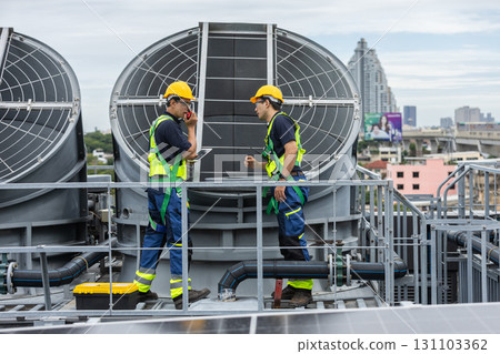 Two engineers in safety gear discuss HVAC system operation while checking data on tablet. Concept of smart facility management, industrial maintenance and mechanical inspection on rooftop. 131103362