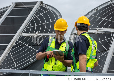Two engineers in safety gear discuss HVAC system operation while checking data on tablet. Concept of smart facility management, industrial maintenance and mechanical inspection on rooftop. Two engineers in safety gear discuss HVAC system operation while checking data on tablet. Concept of smart facility management, industrial maintenance and mechanical inspection on rooftop. 131103363