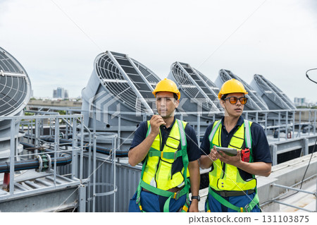 Engineers wearing safety vests and helmets discuss HVAC system inspection on rooftop. Industrial maintenance and building ventilation system operation concept. 131103875