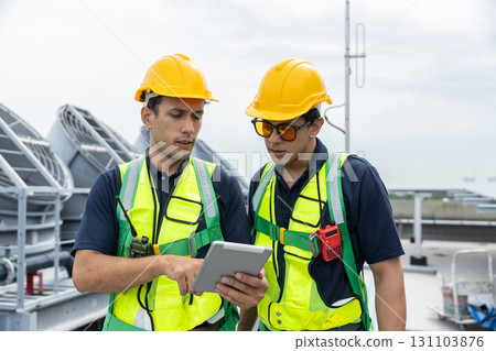 Engineers wearing safety vests and helmets discuss HVAC system inspection on rooftop. Industrial maintenance and building ventilation system operation concept. 131103876