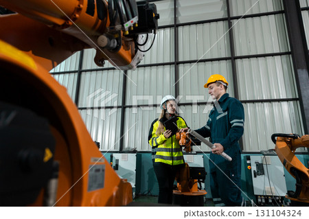 Two engineers wearing safety helmets and reflective workwear discussing project with tablet inside factory, surrounded by robotic arms. Concept of automation, teamwork, smart industry, and safety. 131104324