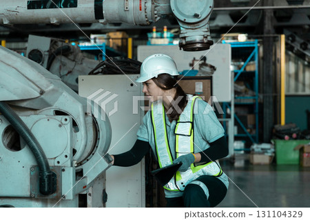Female robot arm engineer wearing safety helmet and reflective vest inspecting industrial machinery inside factory use tablet. Concept of workplace safety, machinery maintenance and smart industry. 131104329