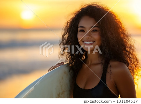 A smiling young woman with curly hair enjoys a beautiful sunset at the beach, holding her surfboard 131104517
