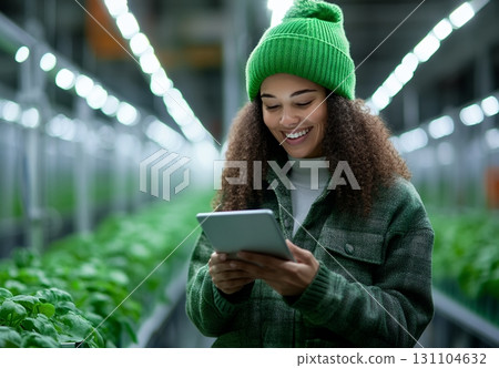 A smiling young woman in a green hat uses a tablet in a modern indoor farm 131104632