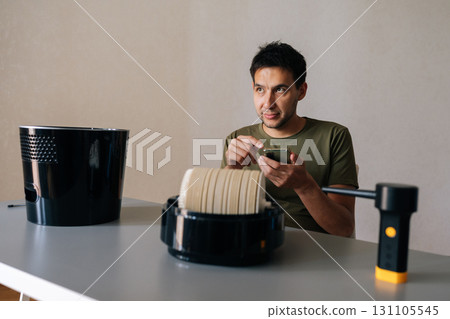 Man sitting at table and cleaning air humidifier filter, using instructions or tutorial on smartphone, with filter, blower and humidifier body visible. Concept of maintenance modern home appliance. Man sitting at table and cleaning air humidifier filter, using instructions or tutorial on smartphone, with filter, blower and humidifier body visible. Concept of maintenance modern home appliance. 131105545