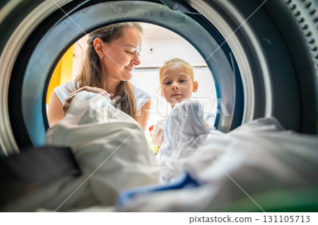 Mother and daughter doing laundry together in a self-service public laundromat, sharing household chores, building responsibility and cooperation in everyday family routine 131105713