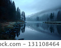 Blue hour mist drifts over a calm mountain lake, pine silhouettes reflected in glassy water, frosty shore 131108464