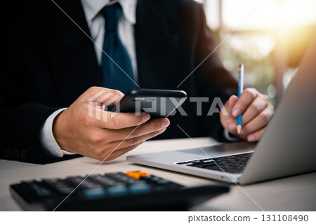 Business person interacting with mobile phone and laptop. Suitable for digital transformation, mobile banking, fintech, online work, and business planning visuals. 131108490