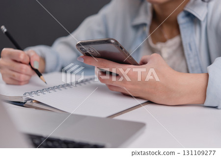 young woman using a smartphone while taking notes, showcasing the balance between digital communication and traditional note-taking in a modern workspace. 131109277