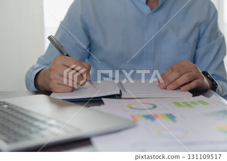 Focused businessman writing down business strategy with financial data charts. Office work environment for analysis and decision making. 131109517