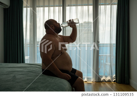 Oversize guy drinks lot of water sits on bed, trying to cool off resist heat cope with dehydration Oversize guy drinks lot of water sits on bed, trying to cool off resist heat cope with dehydration 131109535