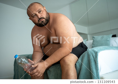 Tired fat man sits on bed with water bottle after training for weight loss think about goal future Tired fat man sits on bed with water bottle after training for weight loss think about goal future 131109543