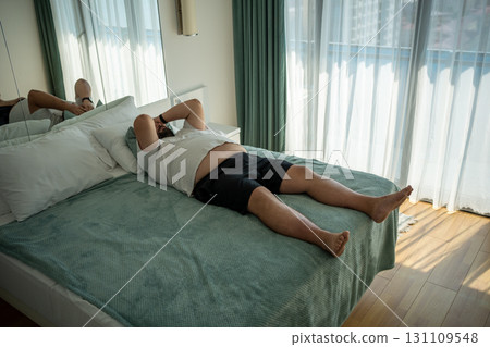 Fat oversize man suffer dying from heat. Lazy, obese overweight guy lies on bed, feels tired in room 131109548