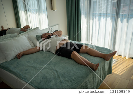Fat oversize man suffer dying from heat. Lazy, obese overweight guy lies on bed, feels tired in room Fat oversize man suffer dying from heat. Lazy, obese overweight guy lies on bed, feels tired in room 131109549