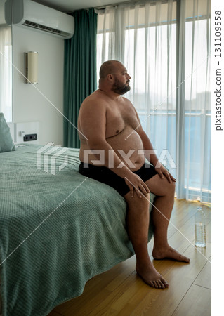 Overweight man in shorts sitting in bed in hotel room looking at window on vacation. 131109558