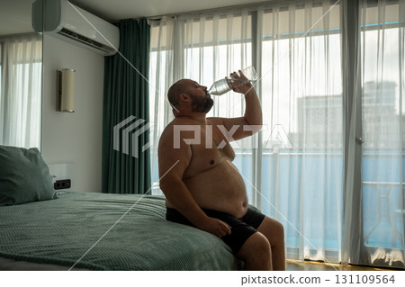 Overweight man drinking water sitting in bed in hotel room on vacation in summer hot weather. 131109564
