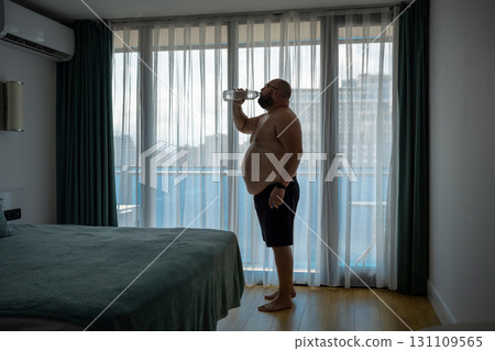 Thirsty overweight man feeling dehydration drinking water in hotel room in hot weather. Thirsty overweight man feeling dehydration drinking water in hotel room in hot weather. 131109565