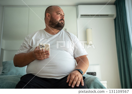 Overweight body positive bearded man with phone and fitness tracker thinking on fatness problem 131109567