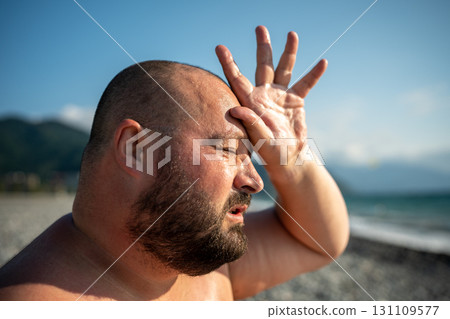 Heavyset man wipes sweat, shields head from sun after heatstroke on beach. Extreme heat under sun Heavyset man wipes sweat, shields head from sun after heatstroke on beach. Extreme heat under sun 131109577