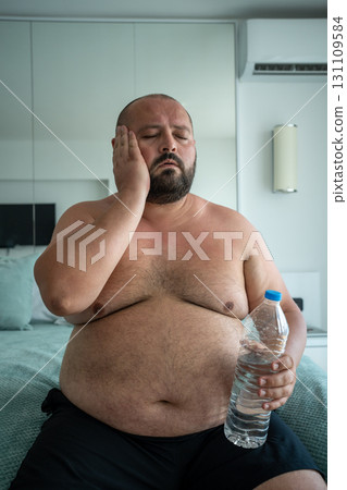 Fat man with water bottle in hand suffering from heat in hotel room on vacation in summer. 131109584