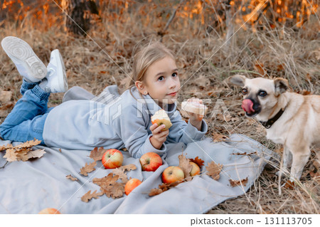 Child lies on a plaid on a picnic, eating apple and give sandwich her dog 131113705