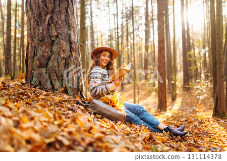 Pretty young woman in hat reading interesting book near tree in autumn forest. Enjoying nature Relax Pretty young woman in hat reading interesting book near tree in autumn forest. Enjoying nature Relax 131114378