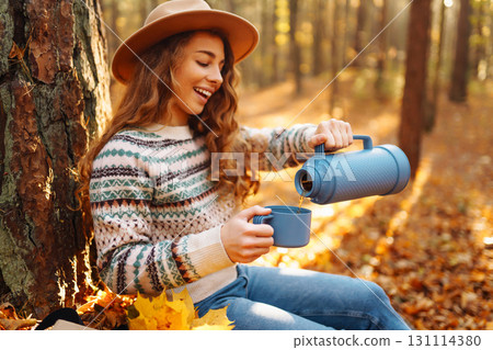 Cute young woman drinks hot drink from thermos and enjoys nature. Autumn landscape. relax concept. Cute young woman drinks hot drink from thermos and enjoys nature. Autumn landscape. relax concept. 131114380