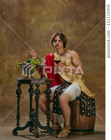 Young man in Roman outfit sitting with wine glass beside grapes and decanter on table Young man in Roman outfit sitting with wine glass beside grapes and decanter on table 131115458