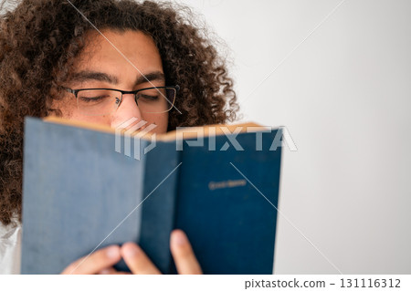 Young man with curly hair and glasses reading a blue book closely Young man with curly hair and glasses reading a blue book closely 131116312