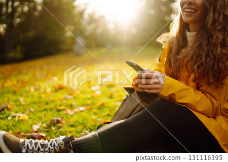 A smiling woman sits outdoors on the lawn among yellow autumn leaves with a phone in her hands. 131116395